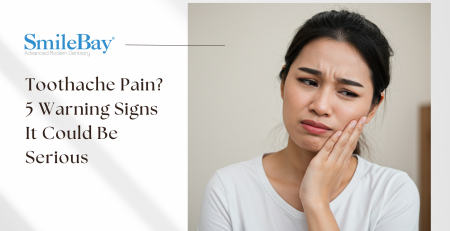 Asian woman with a painful expression holding her cheek, showing signs of a toothache – possible dental issue highlighted by SmileBay Dental.